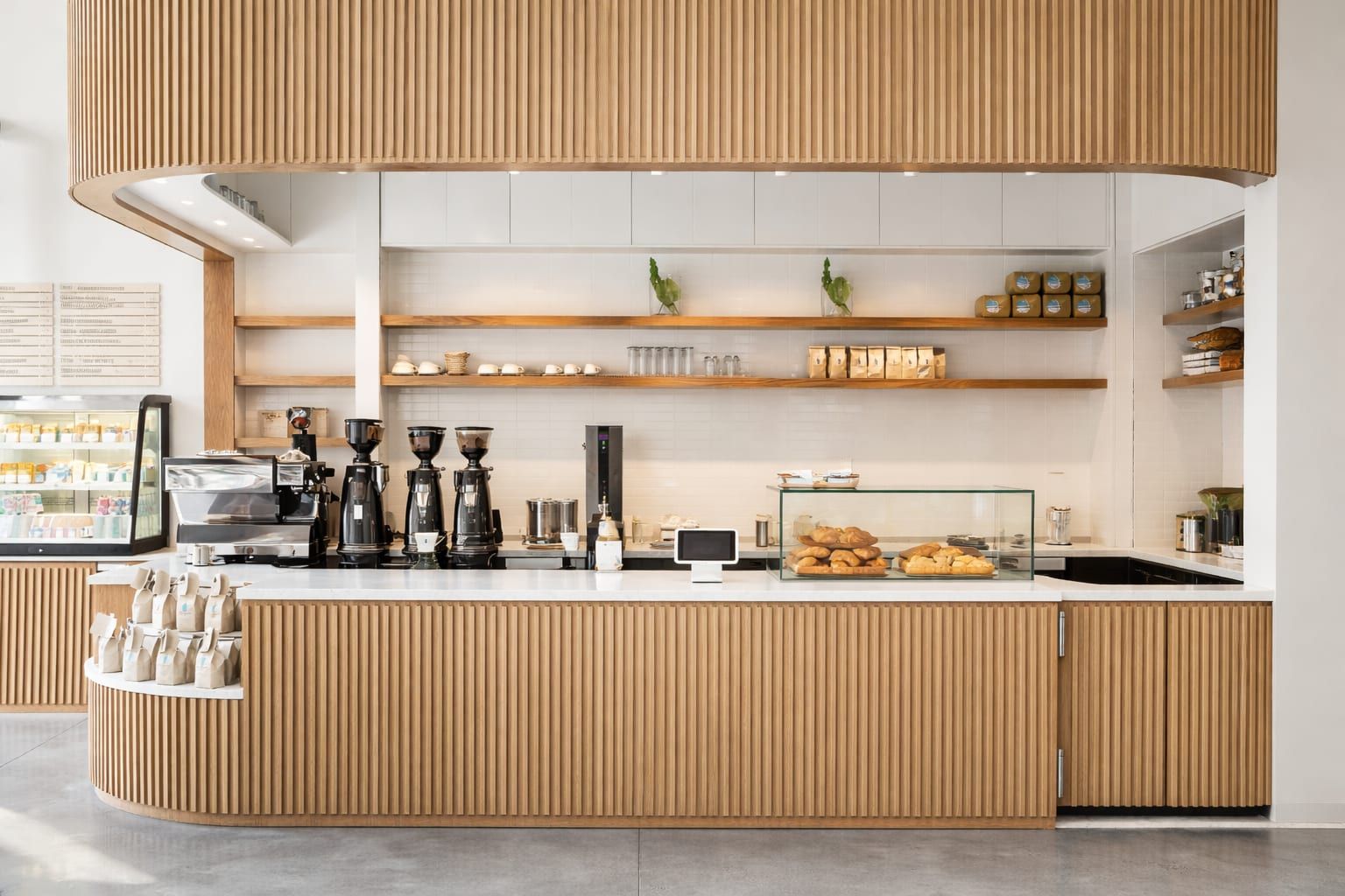Finished Blue Bottle Coffee service counter in San Francisco: curved fluted white-oak counter and matching overhead soffit, open oak shelving on white walls, espresso machines and a glass pastry case