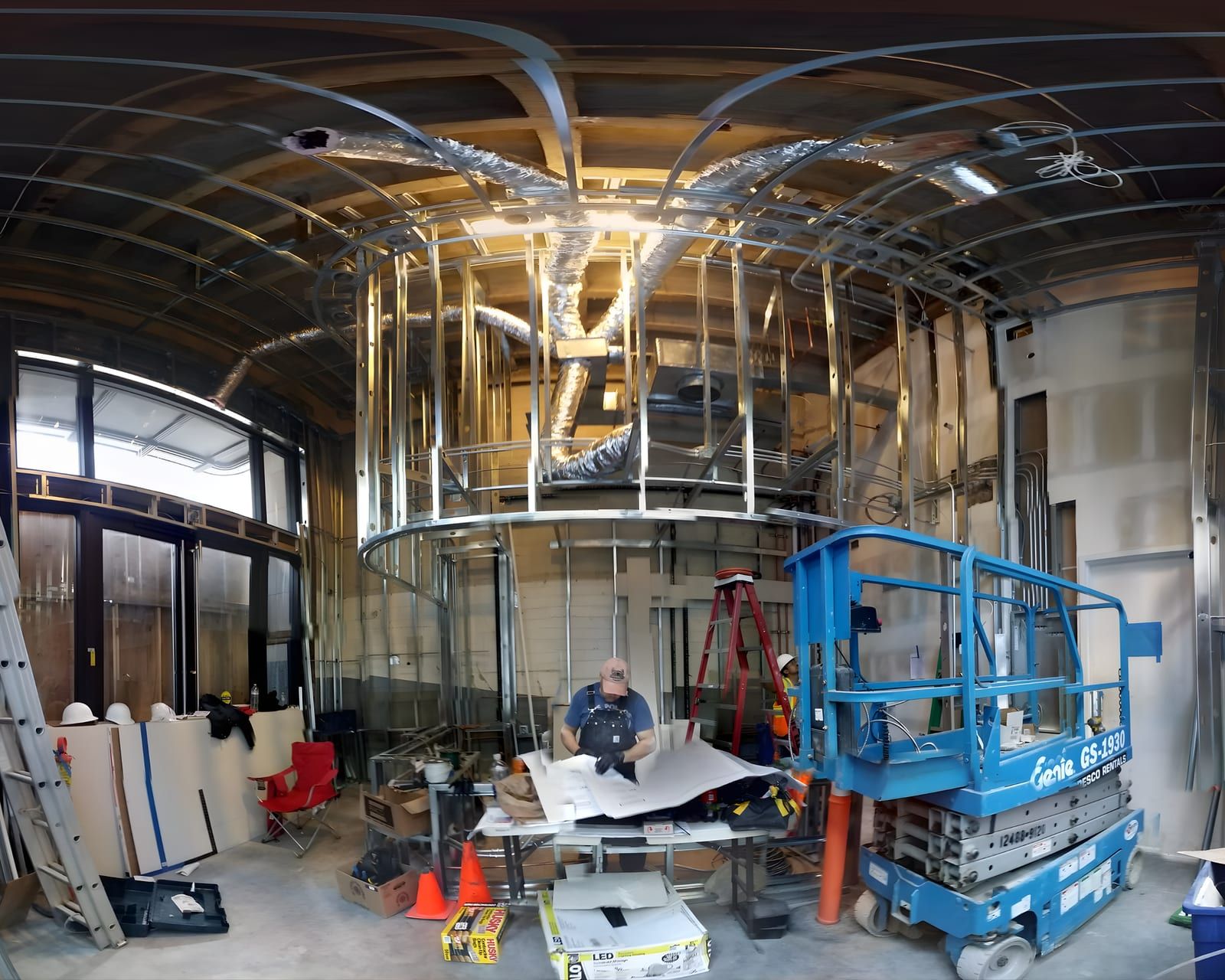 During construction: the curved soffit framed in metal stud with ductwork above, storefront glazing installed, and the crew working from a scissor lift inside the shell